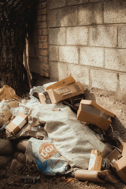 A collection of overflowing rubbish bins and scattered waste bags situated on a paved area in front of a commercial building. The bins include a large grey mixed waste container with a partially open